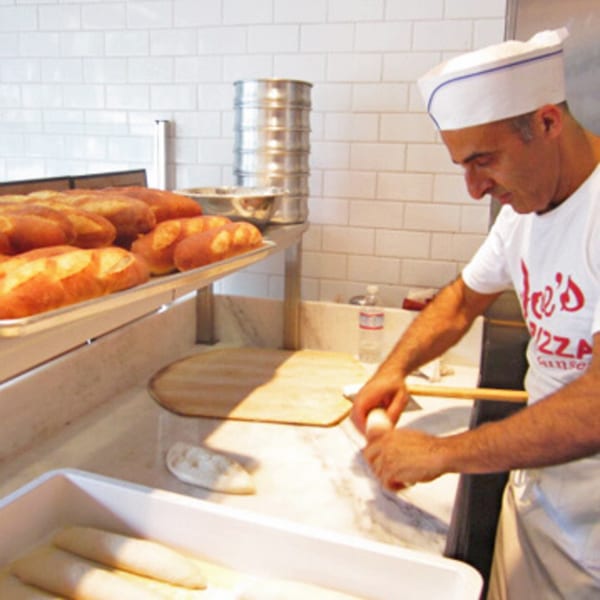 A Joe's Pizza chef in a white hat and shirt prepares dough next to trays of baked bread.