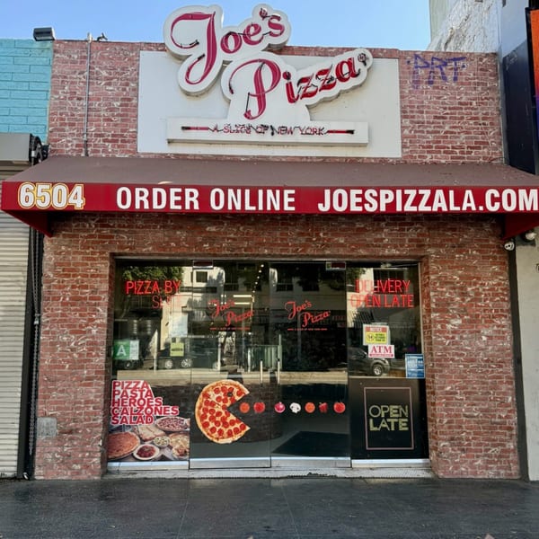 Exterior of Joe's Pizza, a brick building with a red awning and neon sign, featuring a Pac-Man pizza graphic.