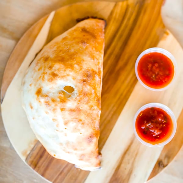 Golden-brown calzone with two cups of red marinara sauce on a wooden board.