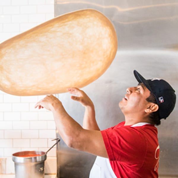 A Joe's Pizza chef in a red shirt and cap expertly tosses pizza dough high in the air.