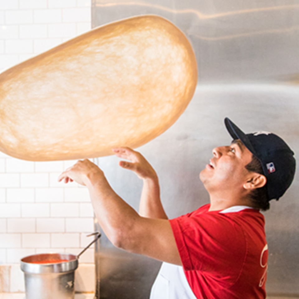 A Joe's Pizza chef in a red shirt and cap expertly tosses pizza dough high in the air.