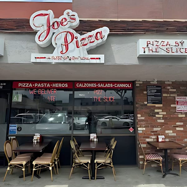 Exterior of Joe's Pizza, featuring a neon sign, menu on the window, and outdoor seating.