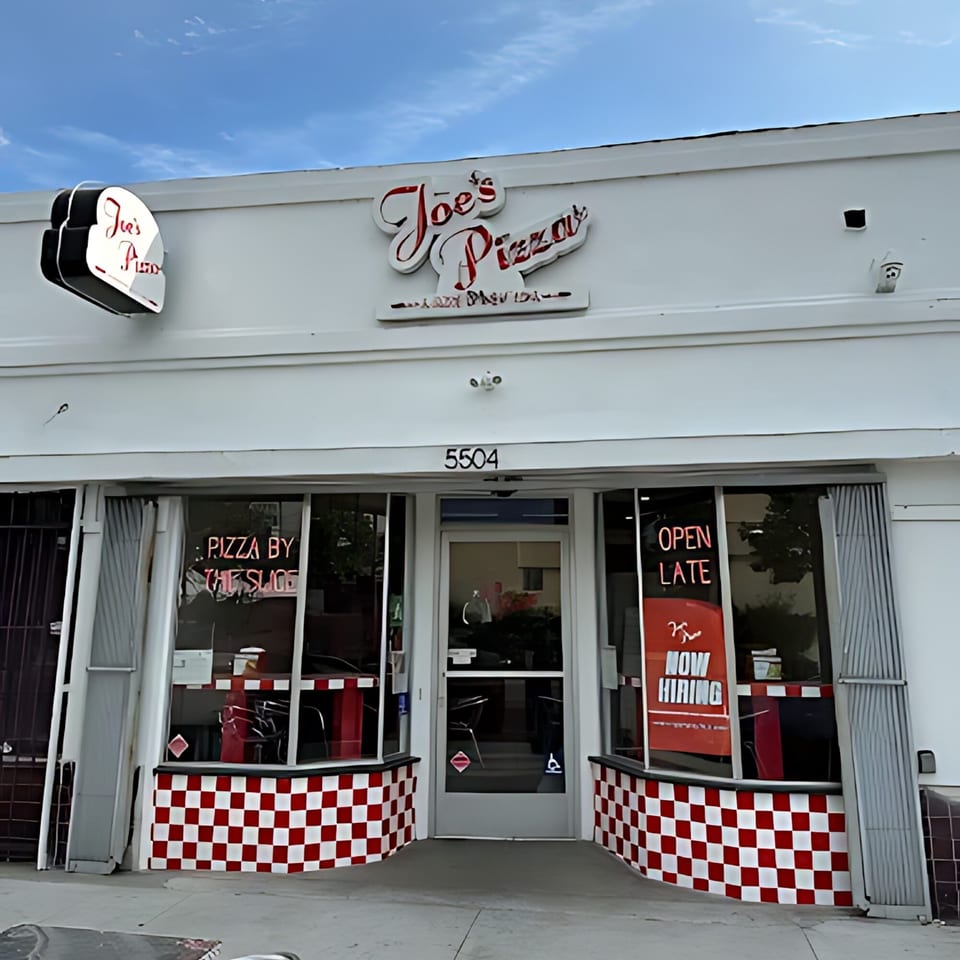 The exterior of Joe's Pizza, a white building with red and white checkered trim, under a blue sky.