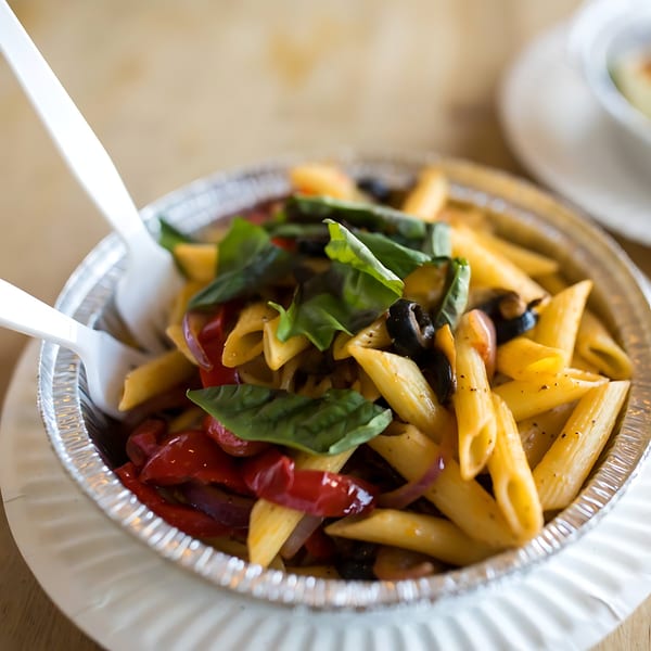 Penne pasta with red peppers, black olives, and fresh basil in a foil container with plastic forks.