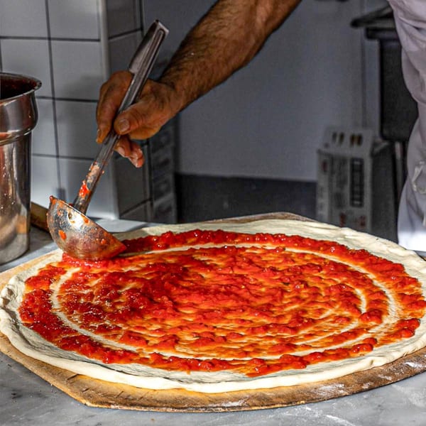 A chef at Joe's Pizza spreads red tomato sauce in a spiral on raw pizza dough.