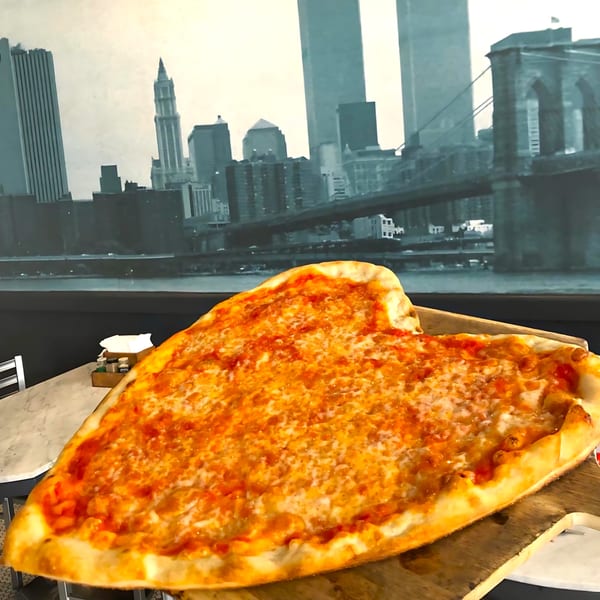 Heart-shaped Joe's pizza with cheese and sauce on a wooden peel, New York City skyline in background.