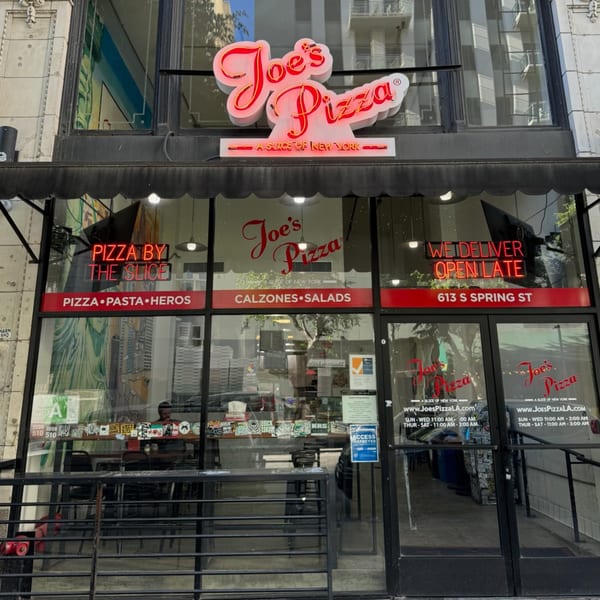 The storefront of Joe's Pizza, featuring a red neon sign and menu items on the windows.