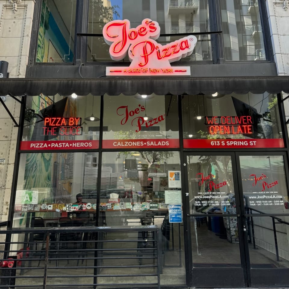 The storefront of Joe's Pizza, featuring a red neon sign and menu items on the windows.