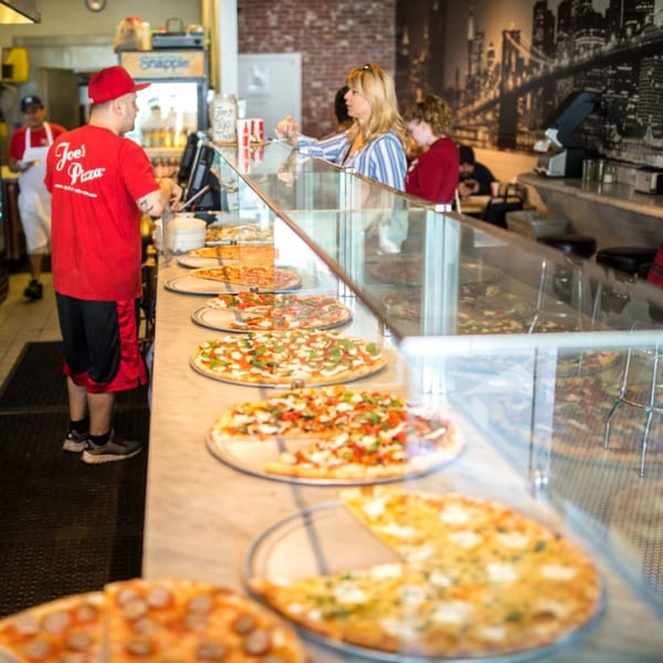 Joe's Pizza counter with a server, customer, and multiple fresh pizzas on display.
