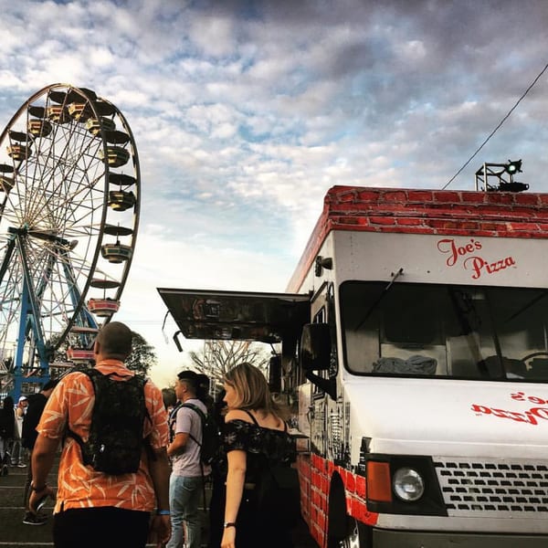 A Joe's Pizza food truck with a Ferris wheel and people in the background under a cloudy sky.