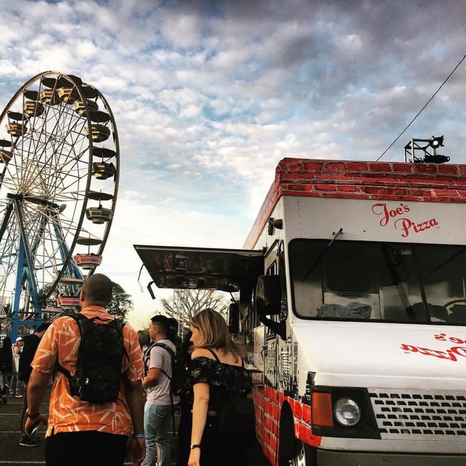 A Joe's Pizza food truck with a Ferris wheel and people in the background under a cloudy sky.