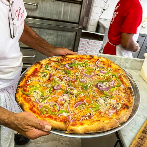 A Joe's Pizza chef holds a freshly baked pizza topped with green peppers, red onions, and mushrooms.