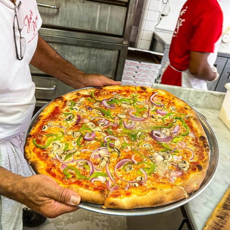 A Joe's Pizza chef holds a freshly baked pizza topped with green peppers, red onions, and mushrooms.