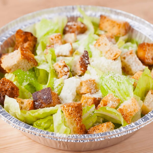 Caesar salad with fresh lettuce, croutons, and grated cheese in a foil takeout container.