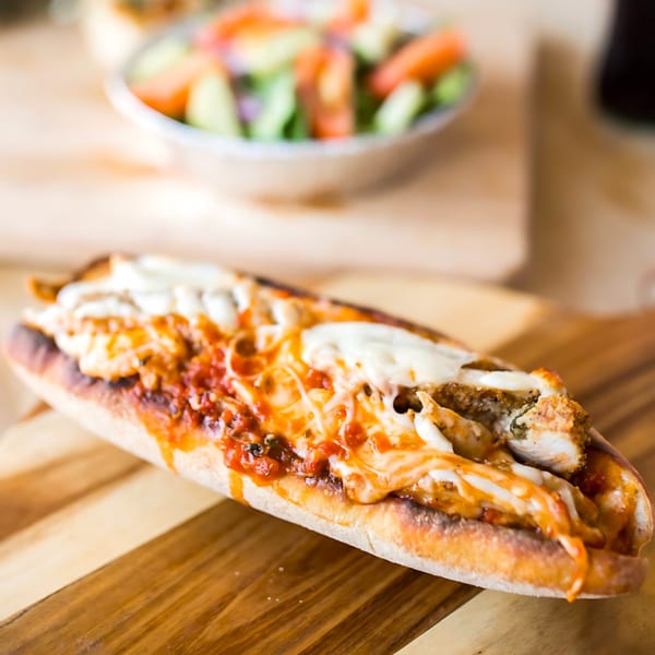 A chicken parmesan hero sandwich with melted cheese and sauce on a wooden board, with a salad in the background.